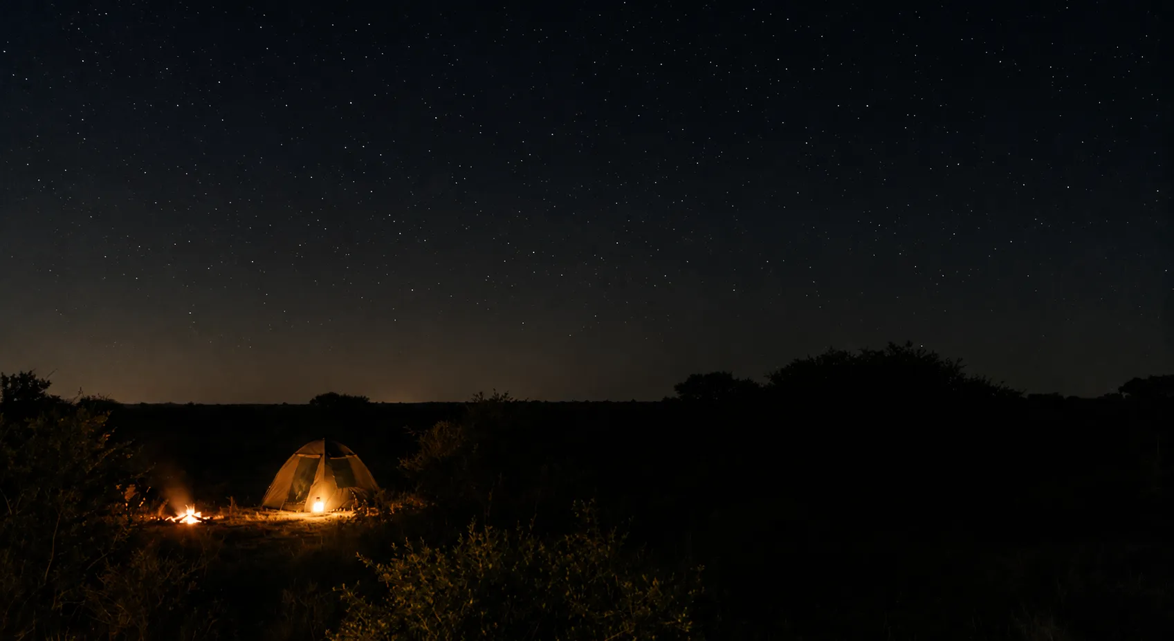 A lit tent and a low campfire at the edge of darkness under a field of African stars.