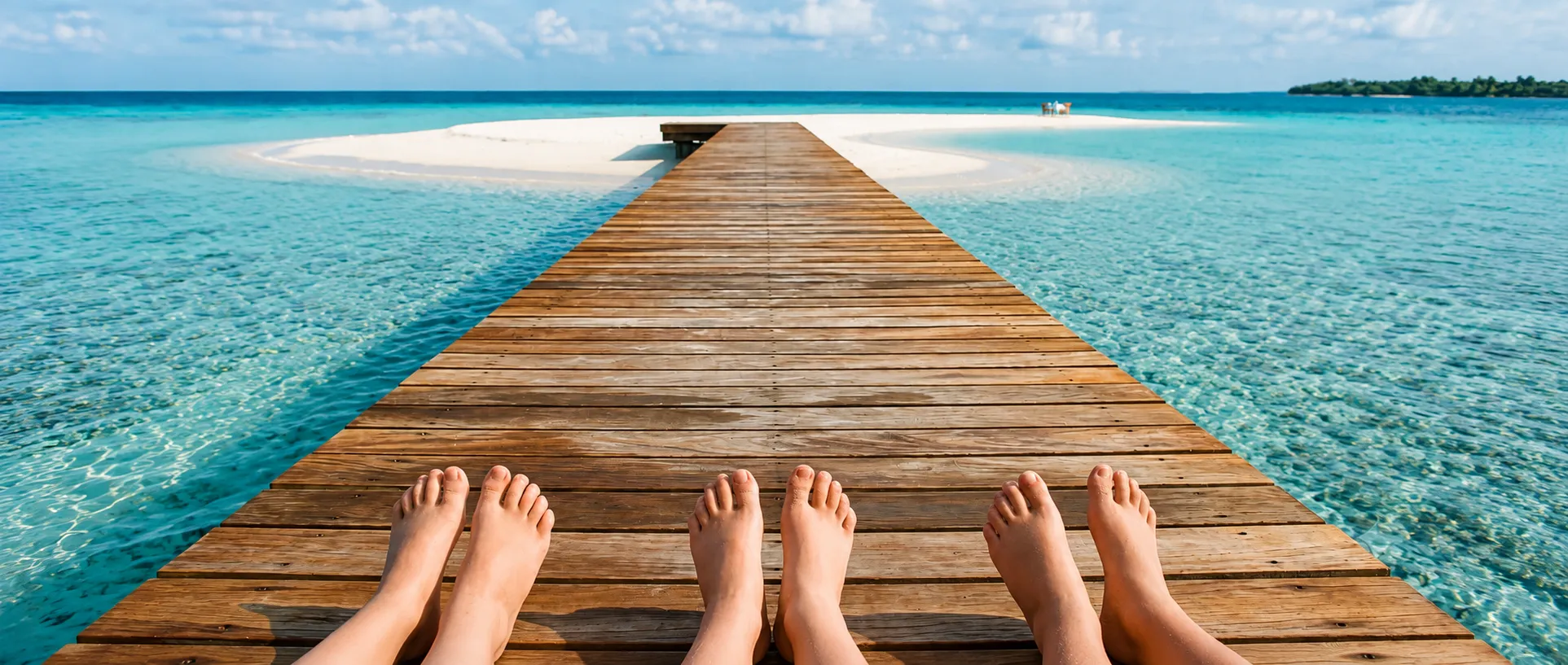 Bare feet on a sunlit jetty above clear lagoon water, a sandbank island beyond.